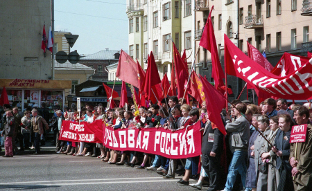 Marchers carry red banners down a street on a sunny day with tall buildings in the background