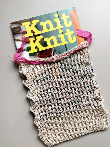 Photo of a knitted bag with a pink ribbon. The bag containing a zine titled Knit Knit.