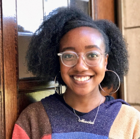 Nicole A. Powell, wearing transparent glasses and large hoop earrings, smiles at the camera