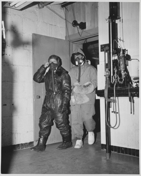 Man in flight suit and man in cold weather gear walking through doorway.