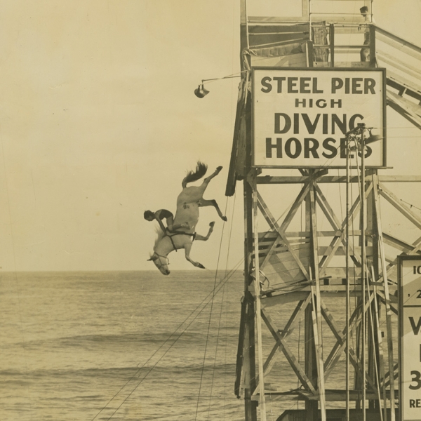 A sepia photograph of Elsa Rahr dressed in equestrian gear mid-dive on a horse from a steel pier high above the ocean water.