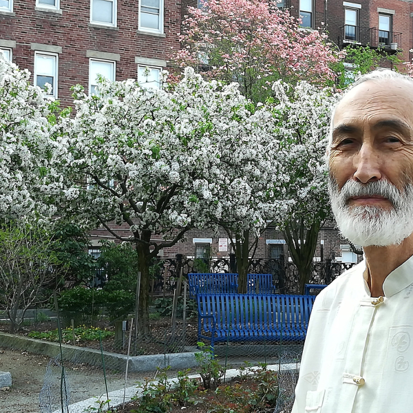 a bearded man in an embroidered shirt standing outside a brick building surrounded by blooming trees