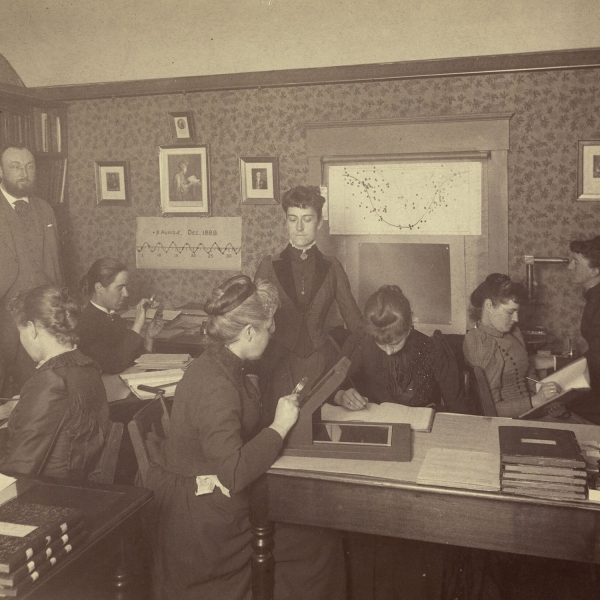 Photo of eight women and one man in a classroom. Maury is seated in profile, third from left, peering into a magnifying glass.