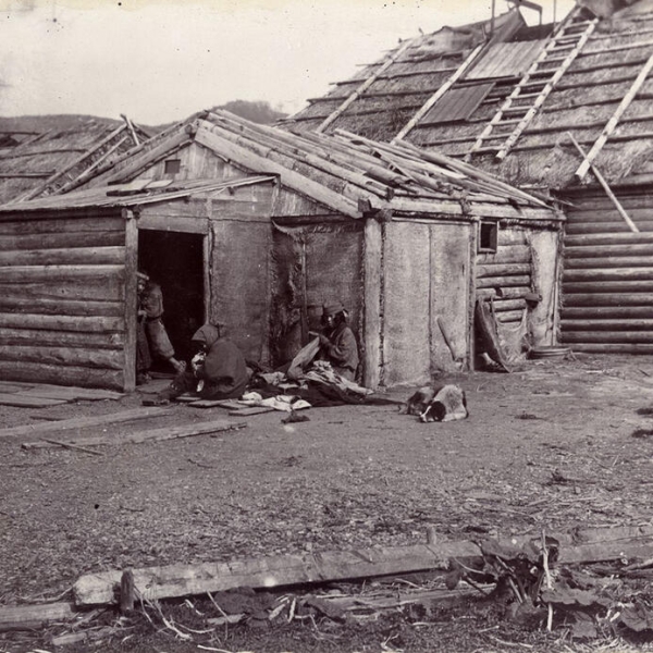 Black and white antique photo of two huts with people sitting in front of them