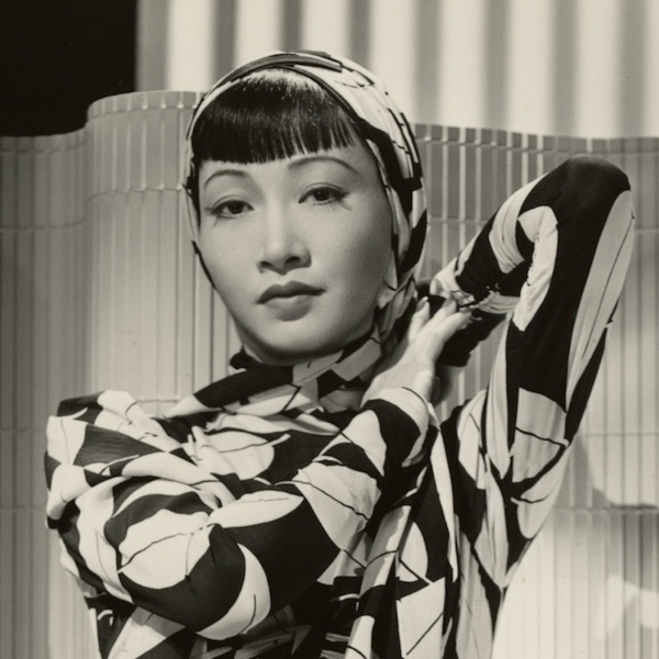 woman in dress with stylized leaves and head scarf poses in front of bamboo screen.