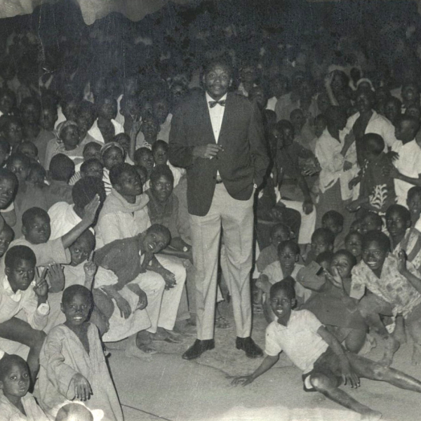 A tall man wearing a suit stands in a room filled with smiling children.