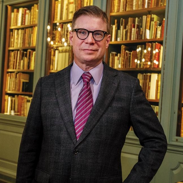 Tom Hyry, wearing a suit, tie, and glasses, stands in front of a bookshelf and looks at the camera