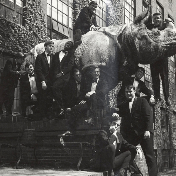 Young men in tuxedos stand on statue of rhino outside of ivy-covered building