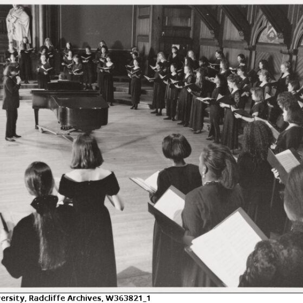 A chorus of several dozen people in floor length black dresses, standing in an arc around a conductor by a grand piano.