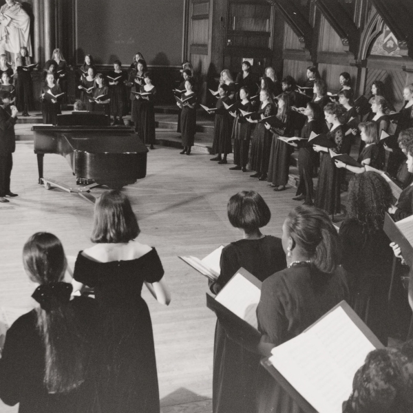 A chorus of several dozen people in floor-length black dresses, standing in an arc around a conductor by a grand piano.