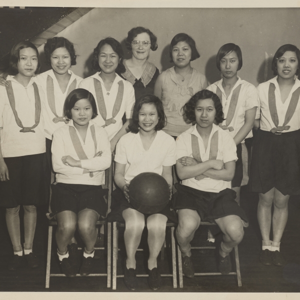 A black and white photograph of the members of a Chinese American girls basketball team, gathered togehter for a team photograph.  The three girls in the front row are seated with the center girl holding a basketball. The other five girls are standing behind them. The girls are in their basketball unifroms, dark skirts and white blouses with a ribbon draped aroud their shoulders. Two teachers or coaches are in the back row center.  