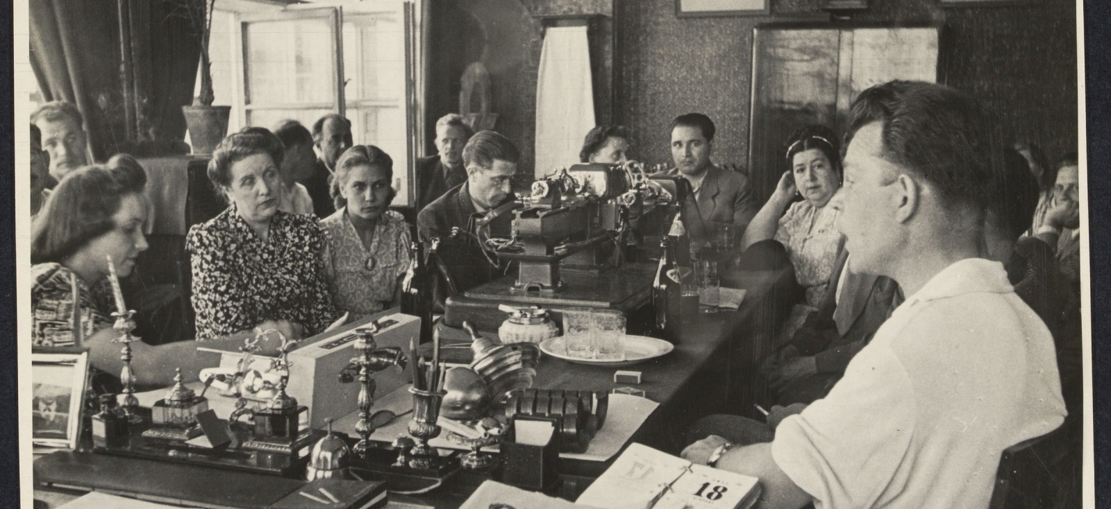 People seated around a table displaying gadgets. Portraits of Lenin and Stalin hang on the wall.
