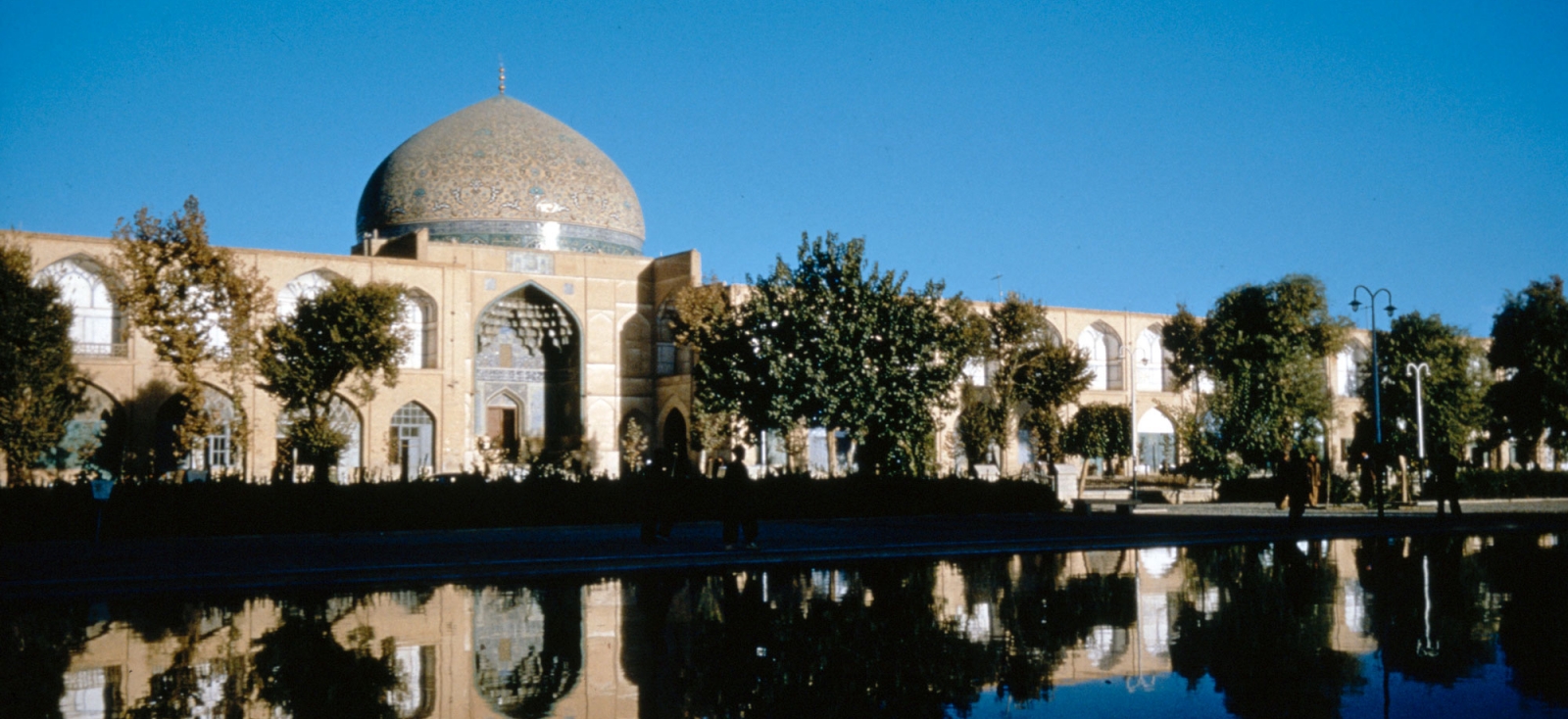 Color photograph showing the Mosque’s portal iwan and dome, both elaborately ornamented with colorful mosaics, and the two-story brick arcade. Their reflection is shown in adjacent tree lined pool in the foreground. 