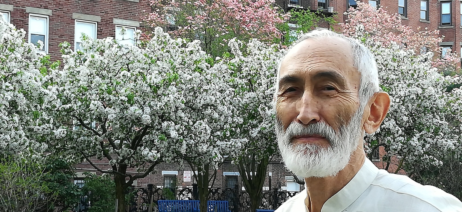a bearded man in an embroidered shirt standing outside a brick building surrounded by blooming trees