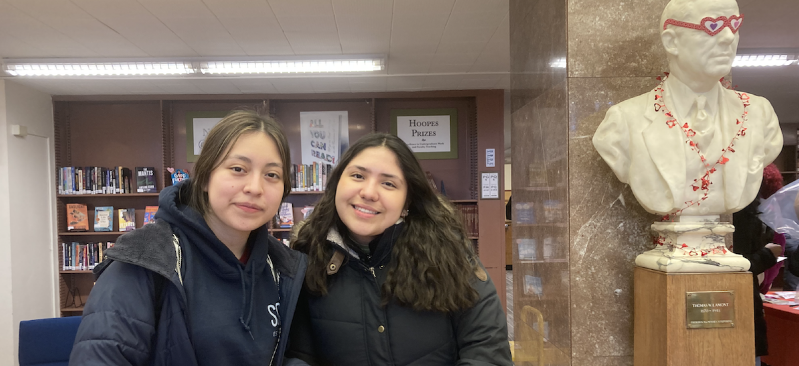 Two students in front of a bust of Thomas Lamont which wears pink heart-shaped glasses