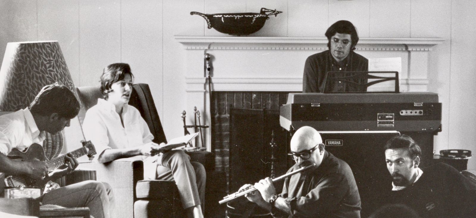 Anne Sexton and Her Kind rehearsing, with a man on the piano, another in the middle playing the flute, and others sitting around in chairs