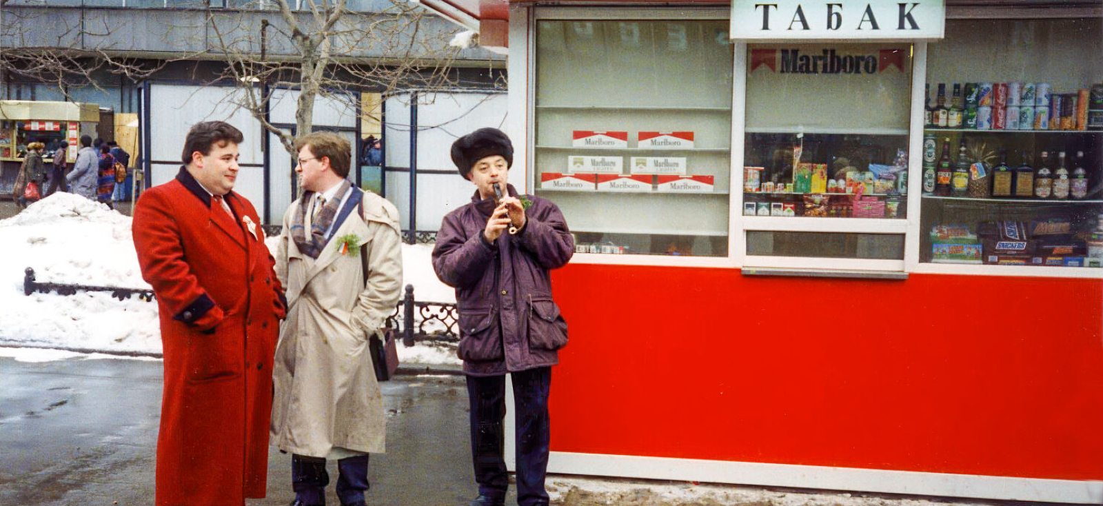 Man in ushanka playing a recorder under a large Marlboro sign; two men stand next to him, one wearing an ostentatious bright-red coat