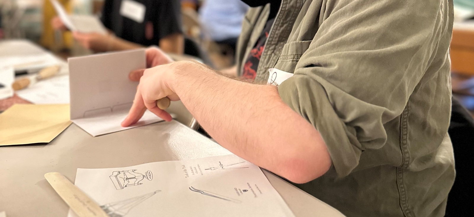 Two people making pamphlets at a table, the one in the foreground with short brown hear and wearing a black face mask, the one in the back with long black hair and without a face mask