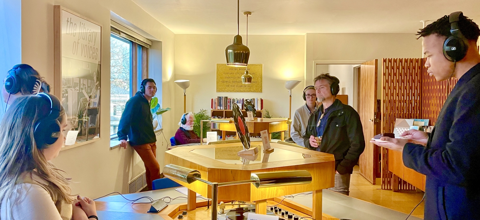 A group of people listen with headphones around a table where a record is displayed on top. A framed photo with The Library of Voices titled on top can be seen clearly on the left wall.