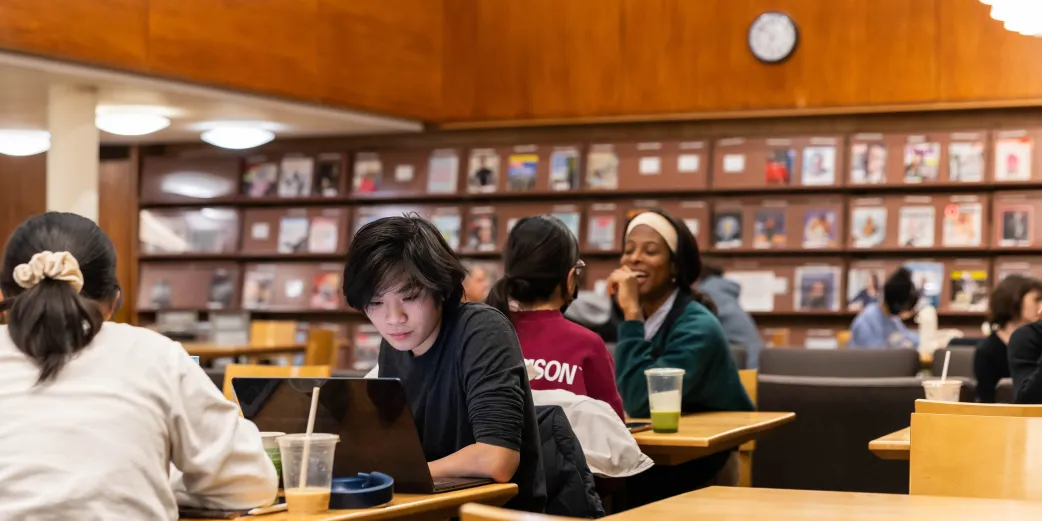 Students chat and work on computers at tables inside a room with books/magazines on display behind them