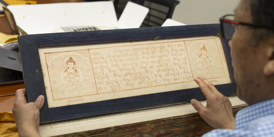 An instructor holds a rectangular Tibetan Buddhist canon block print, featuring text and images.