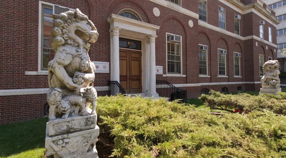 Exterior entrance of the Harvard-Yenching Library, a red brick building with a columned doorway, flanked by stone guardian lion statues and landscaped greenery in the foreground.