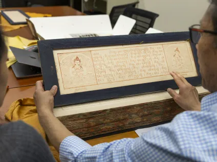 A man his holding up a blockprint page from the Tibetan Buddhist canon. He and his student are at a large reading room table, and looking at the canon. The page has two deity illustrations on the right and left, and the center is Tibetan text from the canon.