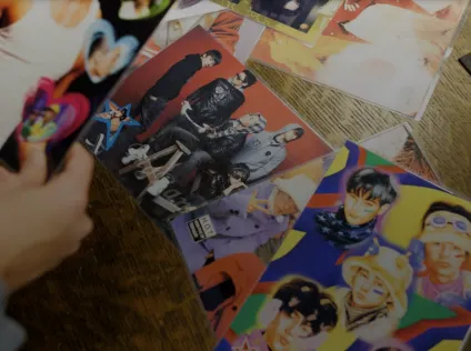 An image of a person looking at a display of k-pop fandom collection items at a table. There's colorful posters on the table and a pair of hands holding a poster.