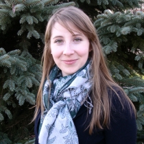 A white woman with light brown hair stands in front of pine trees
