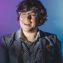 A photograph of Christine Eslao, wearing a grey blazer with a pocket square, laughing against a purple and blue background.