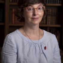 A woman with brown hair and glasses is sitting in front of a case of books. She is wearing a blue blouse with a red poppy pin. 