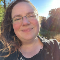 White woman with dark brown long hair wearing glasses smiling while outdoors on a sunny day