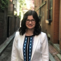 A young woman with shoulder length brown hair and glasses, wearing a white jacket over a dress