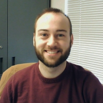 Alex sits in a chair smiling at the camera. Alex has short brown hair and a full beard. He is wearing a red shirt.