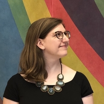 Chelcie smiling and looking up and to the right in front of a rainbow background (a Sol LeWitt wall drawing).