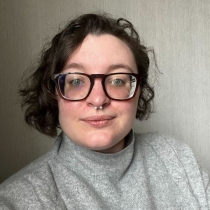 A white, nonbinary person with dark chin length hair, glasses, and grey turtleneck smiles at the camera.