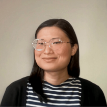 A pale woman with long black hair. She wears round metal-framed glasses.