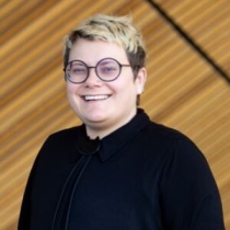 Person with short hair and round glasses wearing a black short-sleeved shirt and bolo tie smiles.