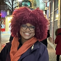 This is a photo of Koukouy Chery smiling. They have a maroon colored afro, an orange scarf wrapped around their neck, and a purple trench coat with a panther brooch. Behind them, the street is lit and people are walking.