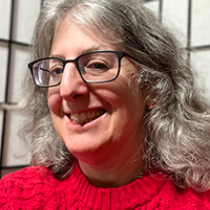 head shot of a woman in her 50s. She has light grey/brown hair about shoulder lenght.  She is wearing black glasses and a red sweater.