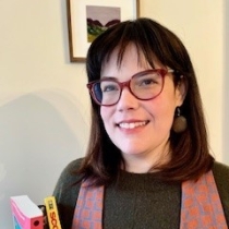 Woman with dark brown hair, light skin, and red glasses holds two books and smiles in front of a small painting of mountains.
