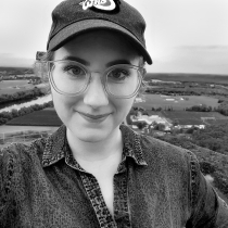 Colleen Cressman wears a baseball cap with her hair pulled back. She has glasses on and is smiling. Behind her in the distance are streams and trees.