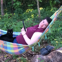 a white woman is lying in a colorful hamnock, holding an e-reader and smiling; a black cat is sitting in her lap