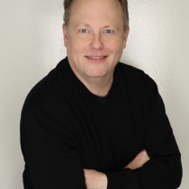 Pale, balding, blue-eyed, brown-haired late-middle aged man in a dark outfit. His arms are crossed. We see him from the waist up.