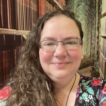 White woman with long wavy brown hair streaked with grays, smiling against a fantasy backdrop of bookshelves set in a forest